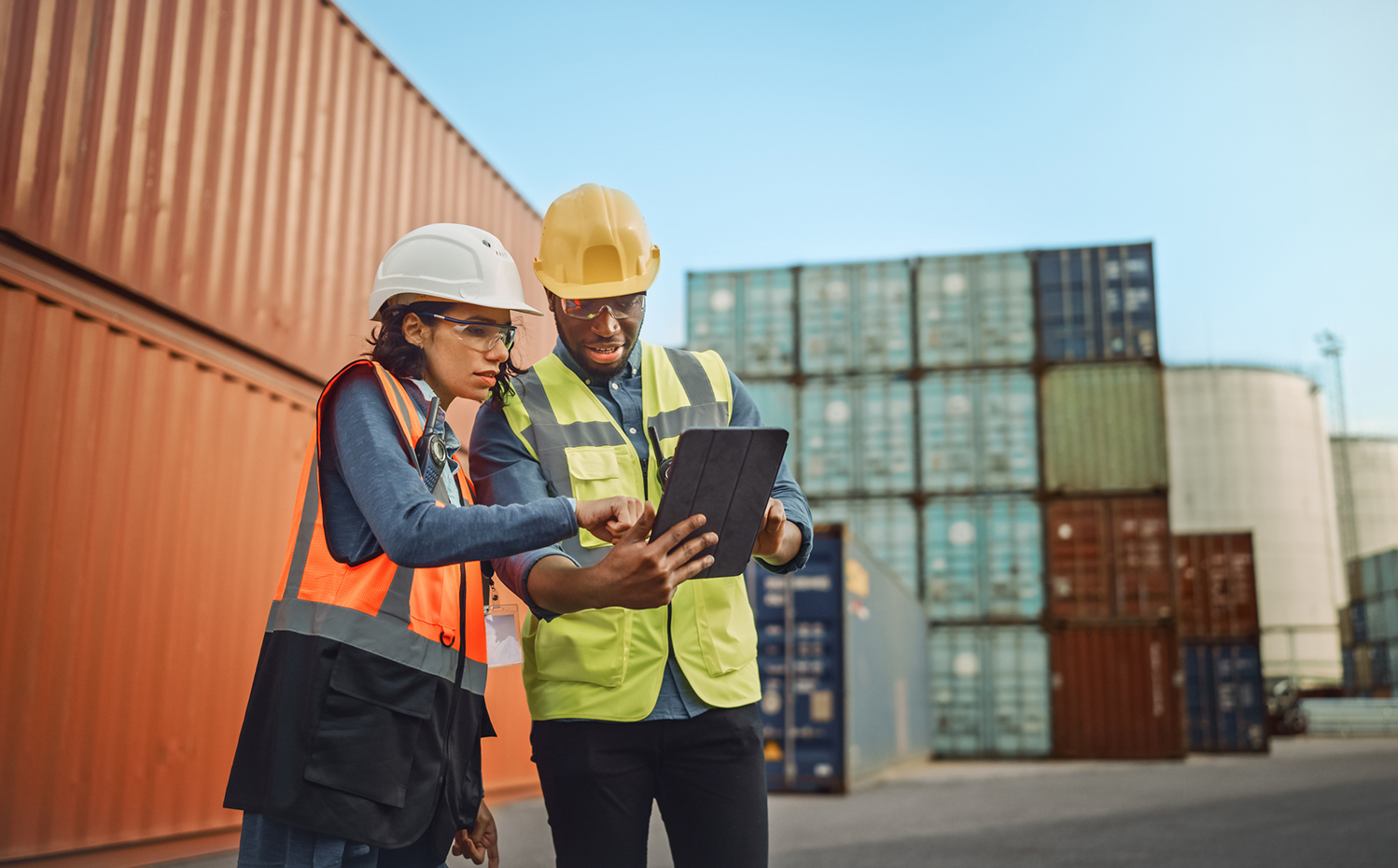 Multiethnic Female Industrial Engineer with Tablet and Black African American Male Supervisor in Hard Hats and Safety Vests Stand in Container Terminal. Colleagues Talk About Logistics Operations.